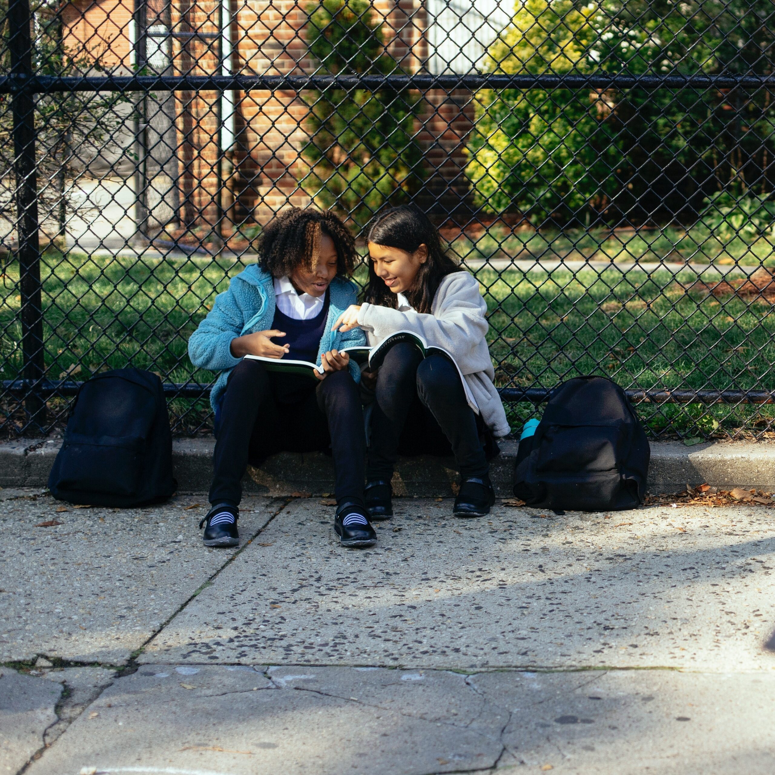 two children with school bags sitting outside and reading and smiling