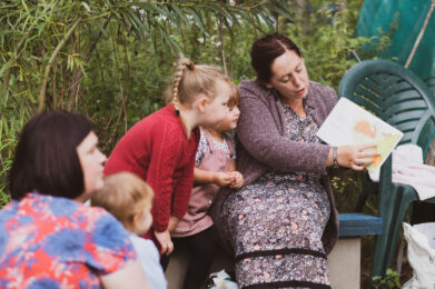 A woman sitting in a green space reading to children from a book, holding the book so the children can see the pictures, and children sitting and standing and looking at the book