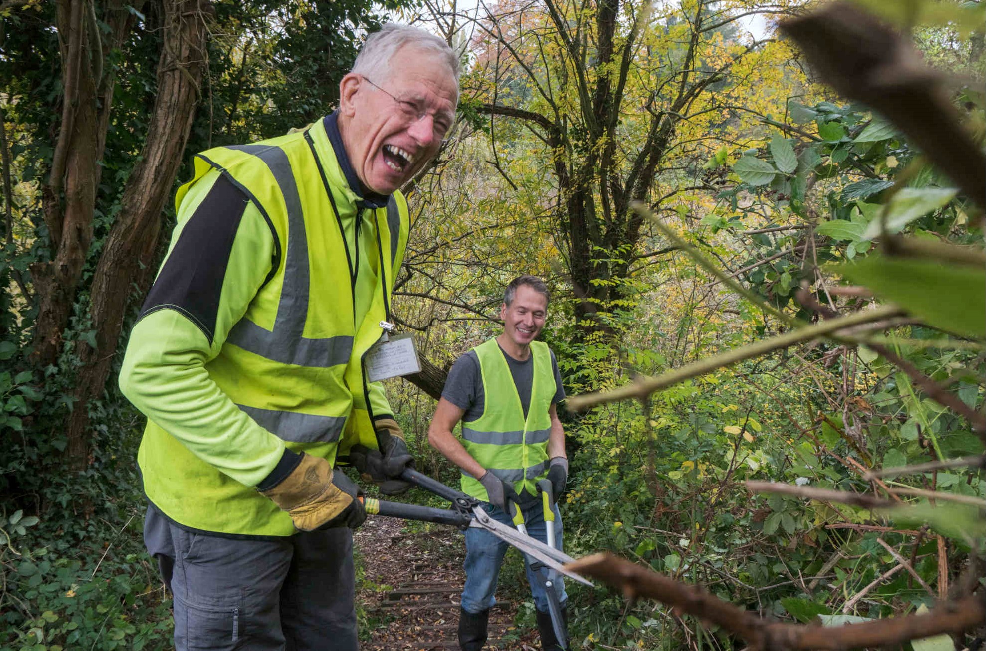 Two people wearing hi vis, standing on a footpath, holding loppers and laughing