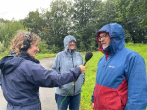 Charlotte Smith smiling and wearing headphones, holding a microphone towards Gareth Roberts, with a third person in the background. All are wearing waterproof jackets. Standing on a path in a green setting in the rain.